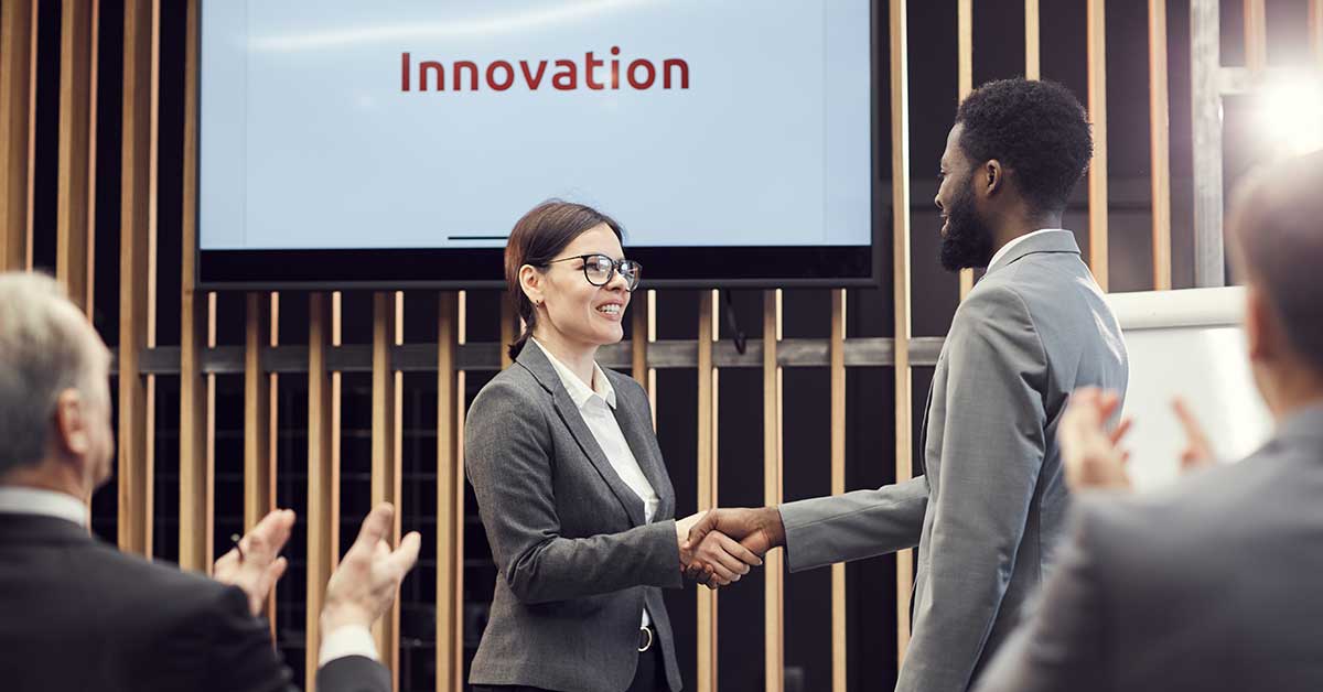 Two business professionals shake hands in front of a screen displaying the word “Innovation” while colleagues applaud in a modern office setting, celebrating the transformative impact of Artificial Intelligence on business success.