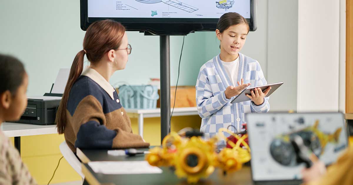 A girl stands presenting with a tablet while two classmates sit and listen; a screen behind her displays a diagram, highlighting the development of future leadership skills in the classroom.