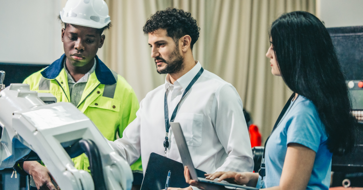 Three workforce professionals stand by a robotic machine, discussing its operation; one wears a hard hat, another holds a clipboard, and the third holds a laptop.