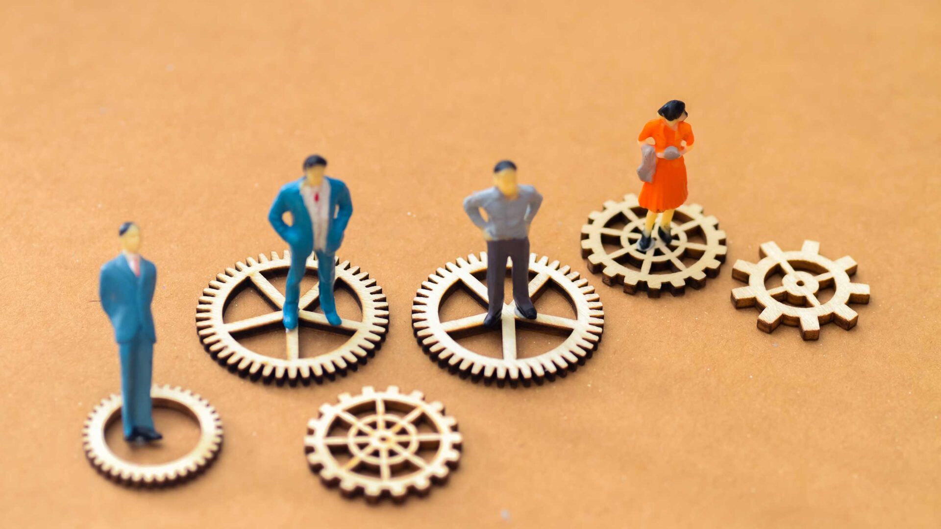 Four miniature figurines, three men and one woman, symbolizing the workforce, stand on interlocking wooden gears placed on a tan surface.