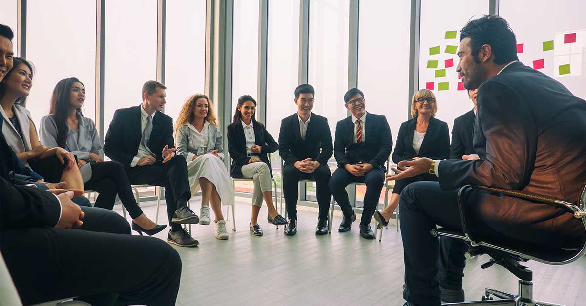 A group of people in business attire sit in a circle having a discussion about leadership in a modern office with large windows, challenging the mindset of “we’ve always done it this way,” that can often be a dangerous sentence for innovation.