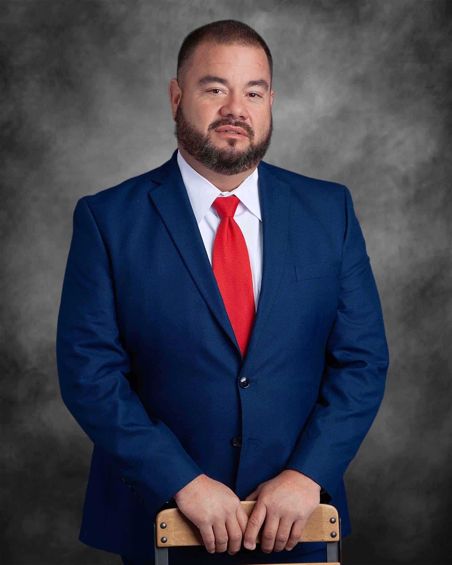 A man in a blue suit with a red tie stands against a gray backdrop, resting his hands on the back of a wooden chair.