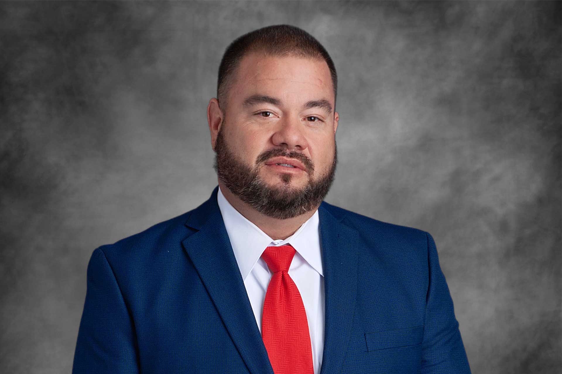 A man with a beard and mustache is wearing a blue suit, white shirt, and red tie, posing in front of a grey studio background.