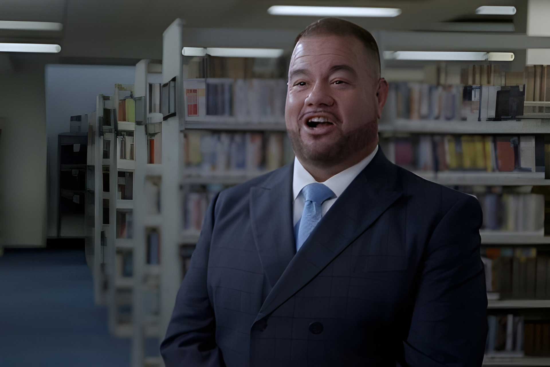 A man in a suit and tie stands in a library with bookshelves in the background. He appears to be speaking or laughing.