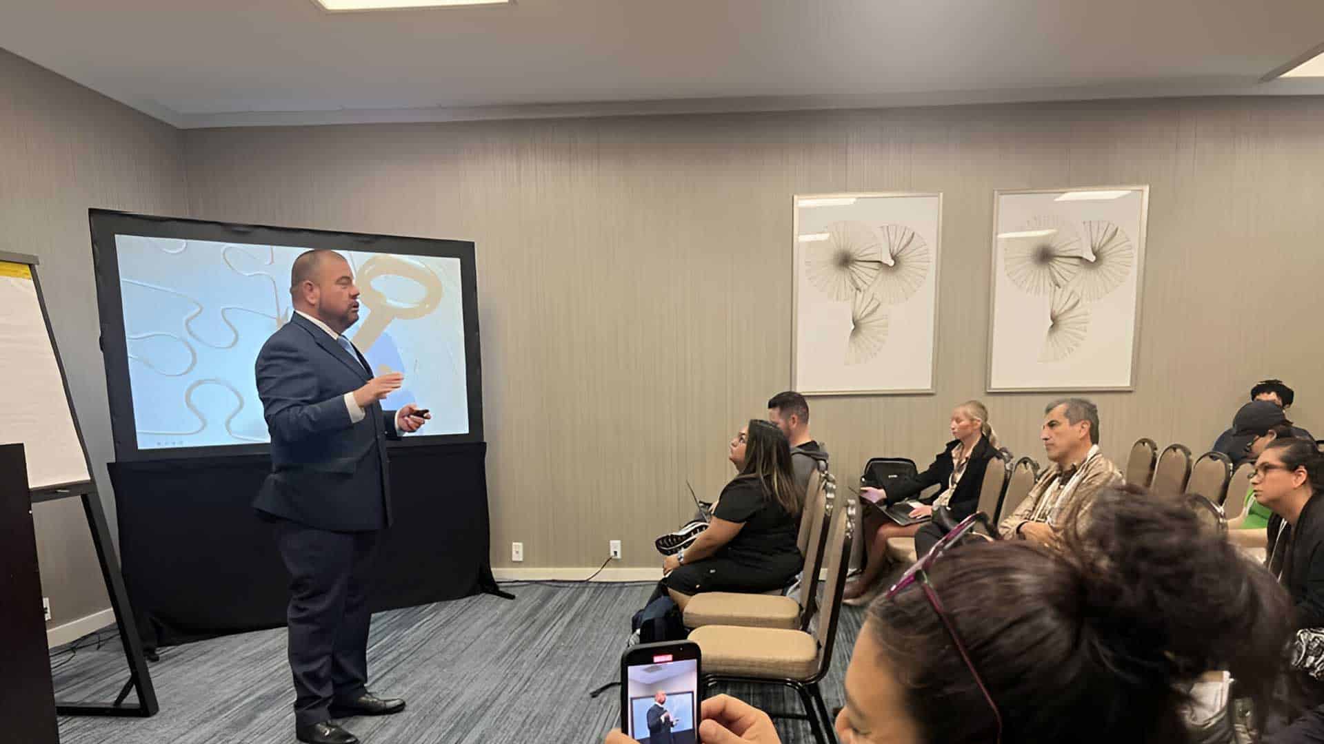 A man in a suit stands in front of a projection screen giving a presentation to a seated audience in a conference room. The audience members are engaged and some take notes or photos.