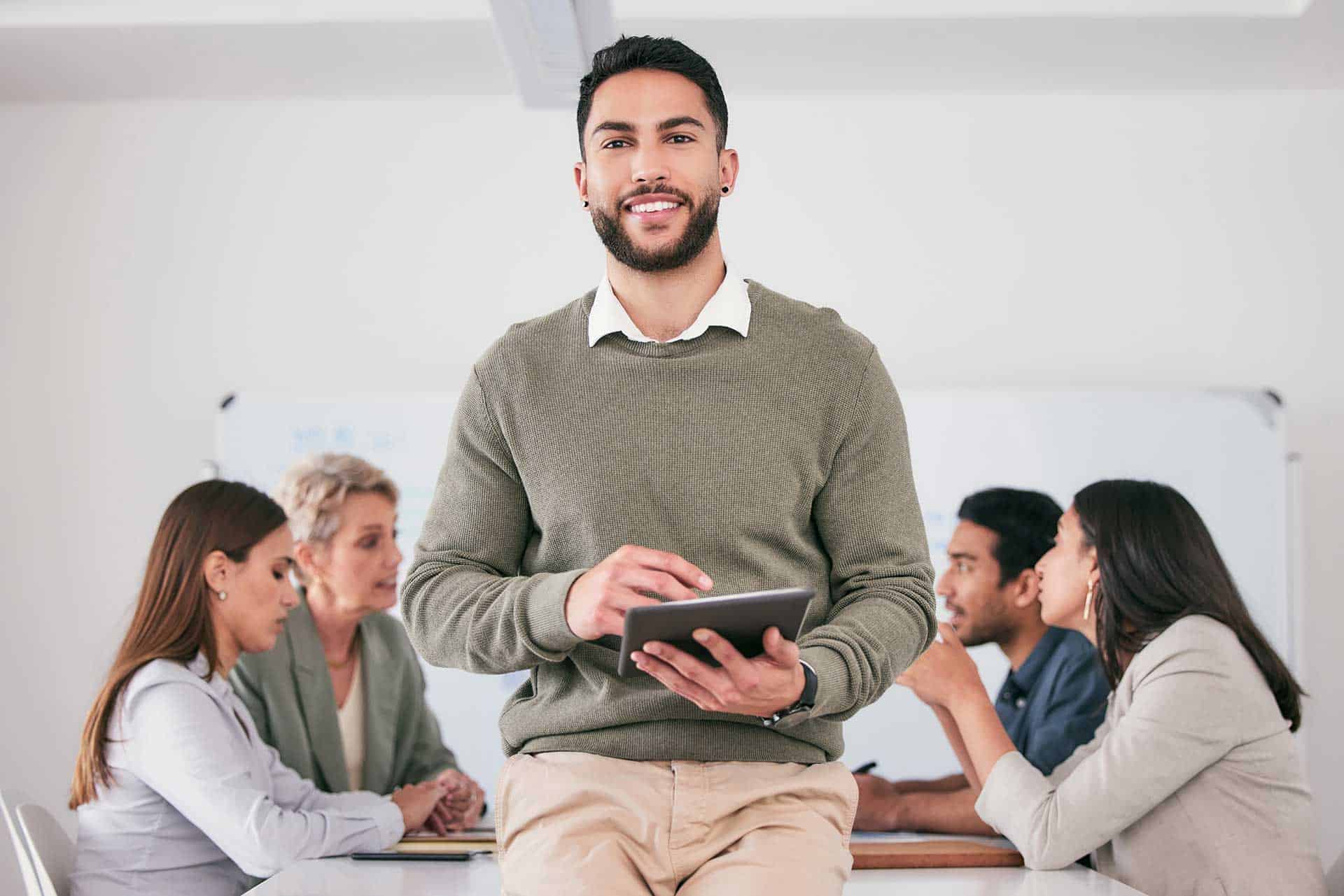 A man stands at the forefront holding a tablet, emphasizing key points on recruitment strategies while four people are seated and engaged in an educational discussion in the background.