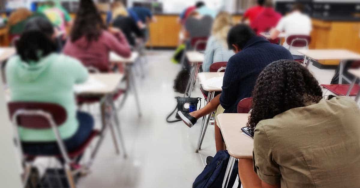 Students sitting at desks in a classroom, focused on their work. A student in the foreground, one of the new immigrant students, has their head down on the desk.