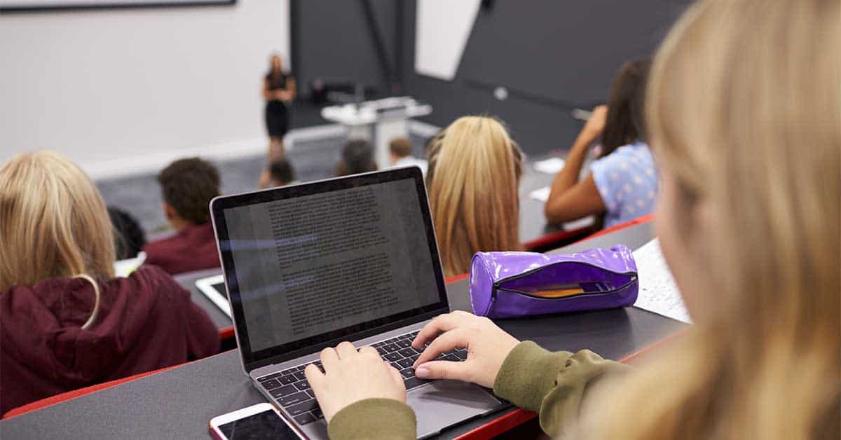 An immigrant student, showcasing bilingualism, types on a laptop while attending a lecture in a large classroom, with other students and the lecturer visible in the background.