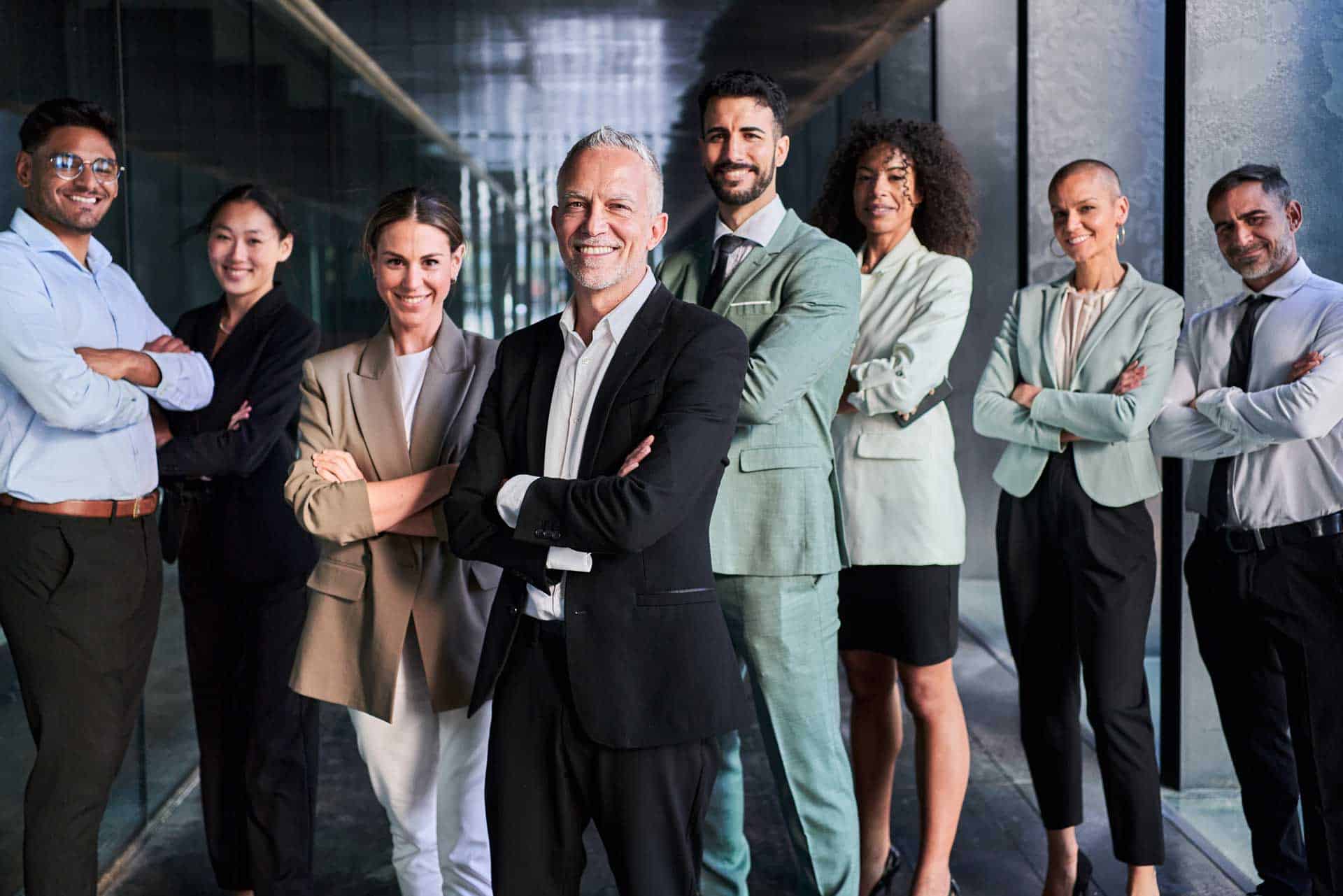 A diverse group of eight professionals stands in a hallway, dressed in business attire, smiling, and posing with arms crossed, showcasing the results of their executive coaching sessions.