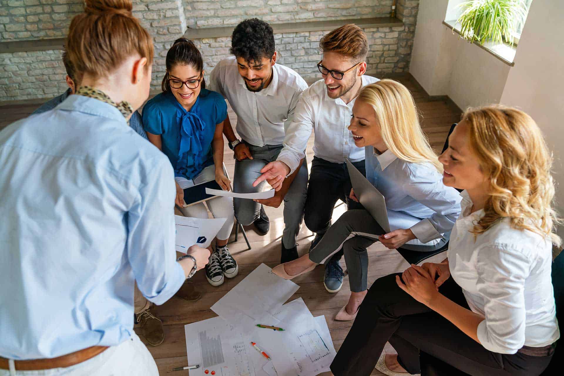 A group of six people, including an executive, sitting and standing in a semi-circle, engaged in a coaching discussion, with papers and diagrams spread on the floor in front of them.