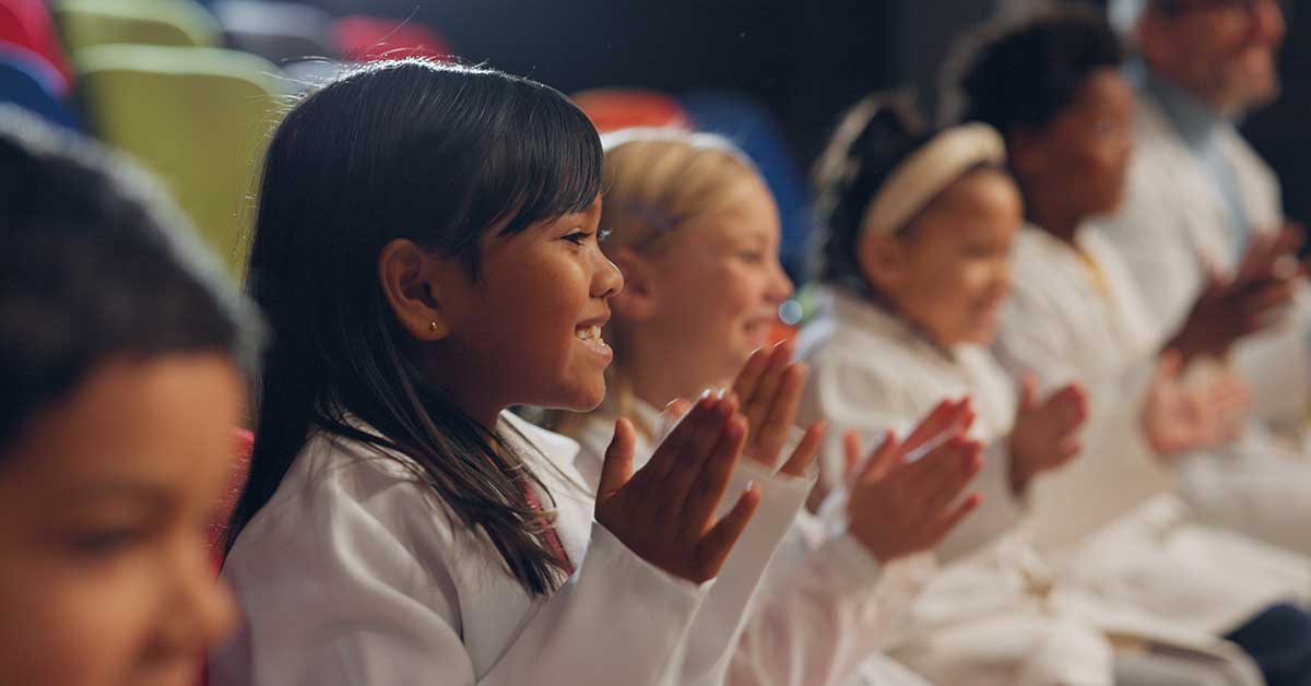 Immigrant students sitting in a row, clapping and smiling, wearing white attire against a background of colorful seats.