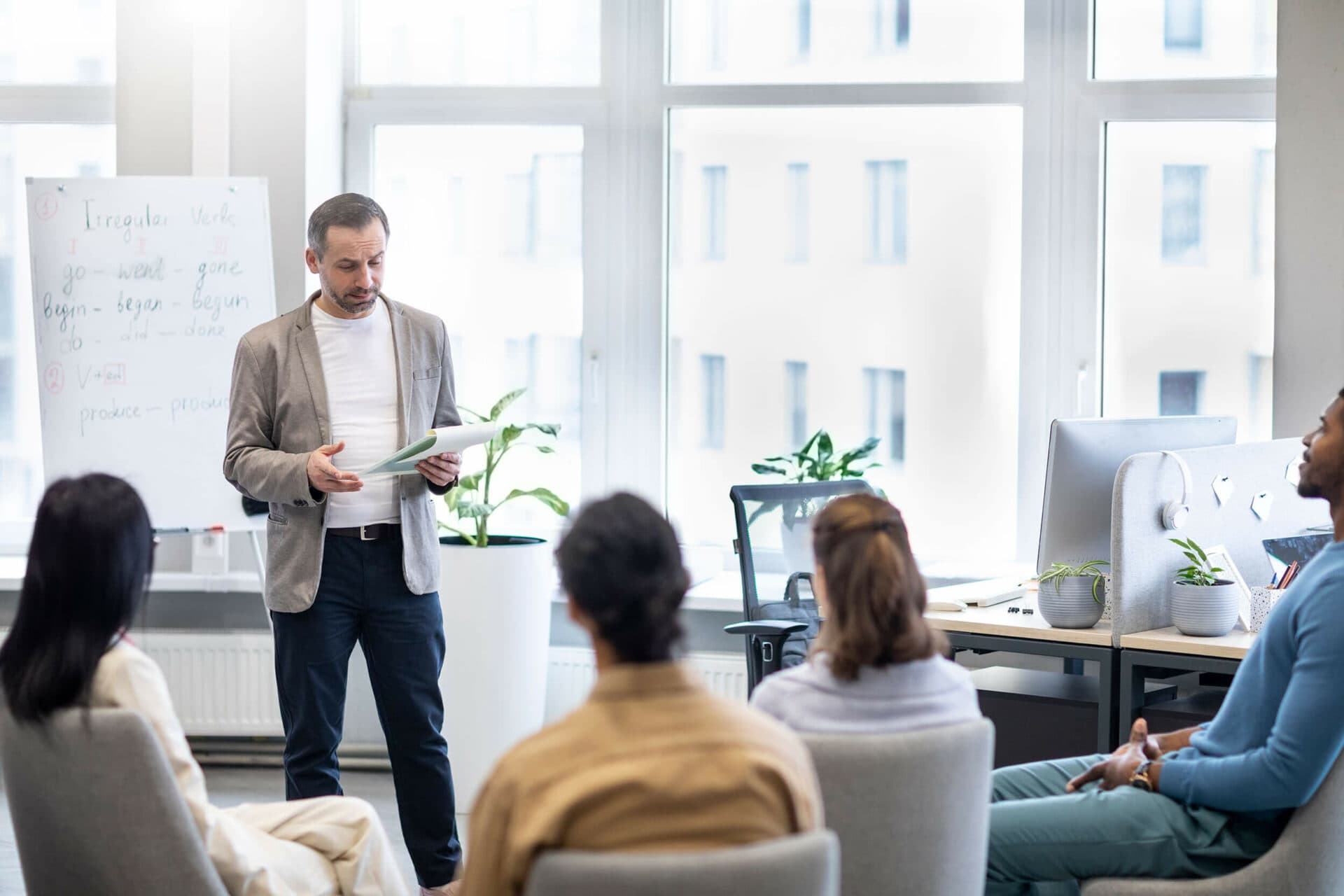 A man stands and speaks to a seated group in a bright office. Behind him, a whiteboard with text is visible, along with large windows and desks equipped with computers and plants.