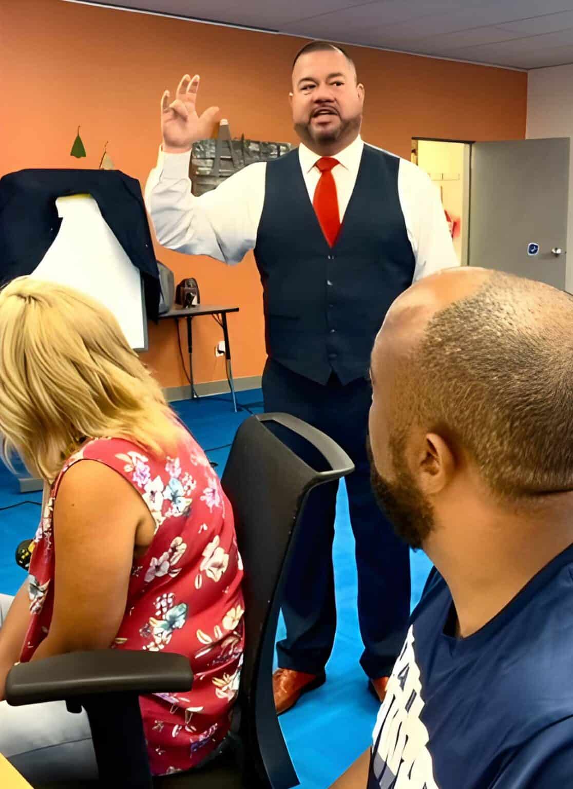 A standing man in a suit gestures while speaking to two seated individuals, a woman with blonde hair and a man with a shaved head, in an office with orange walls and blue flooring.