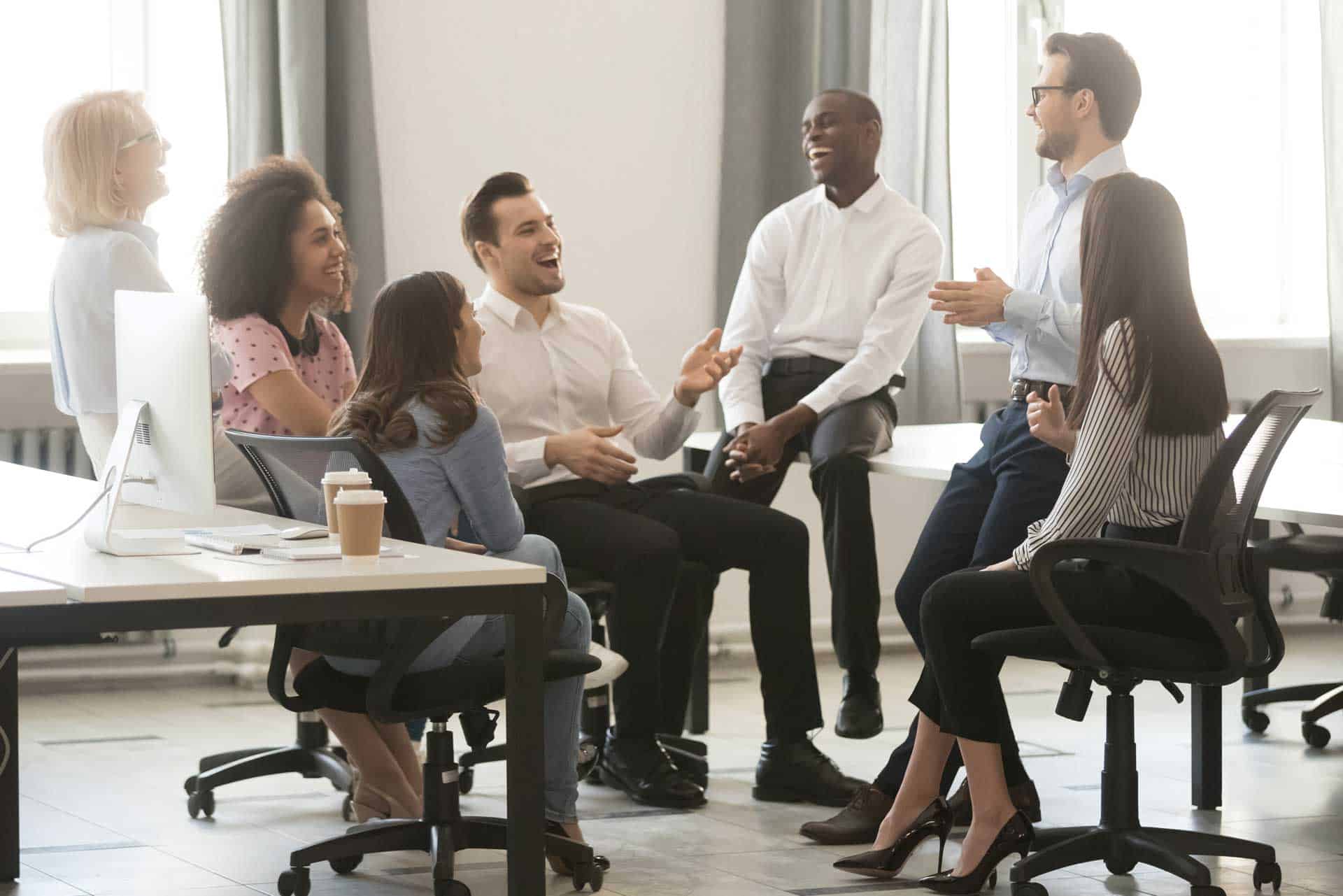 A group of seven people, dressed in business casual attire, are sitting and standing in a brightly lit office, engaged in a lively conversation about executive coaching and laughing.